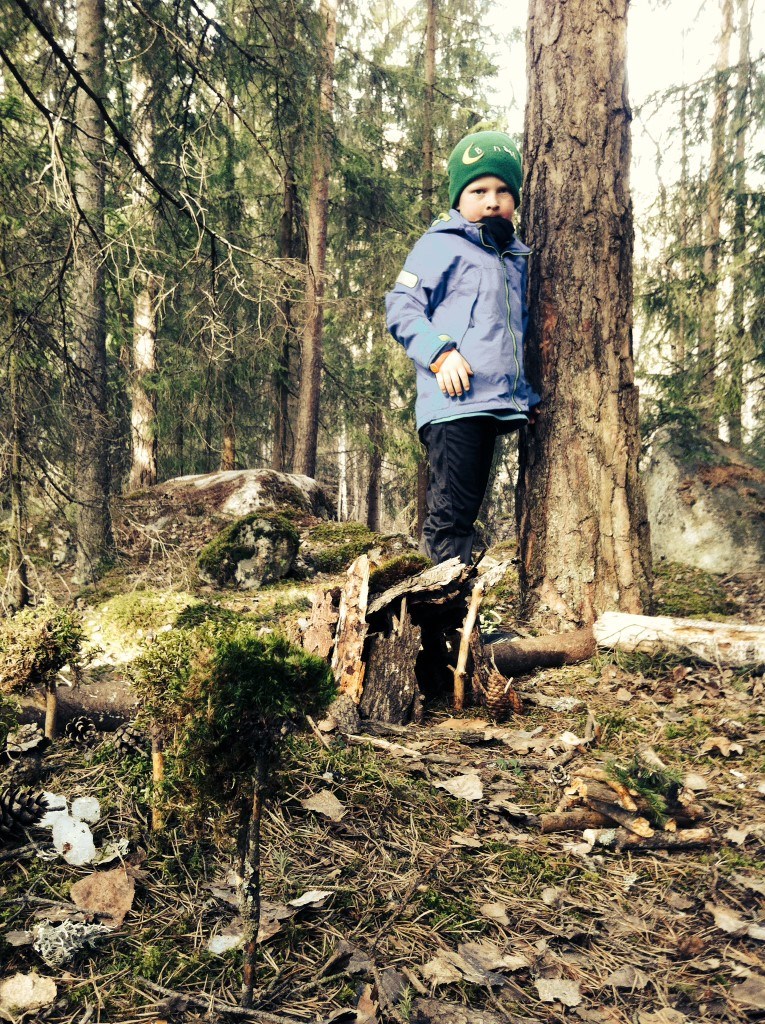 Stolt gutt foran trær, bål og hytte lagd av bark.